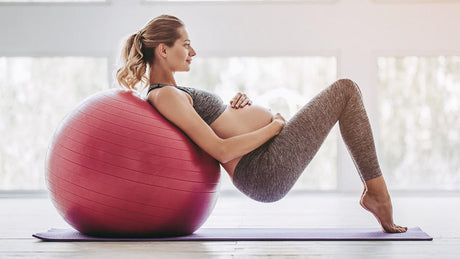 Zwangere vrouw leunt achterover op fitnessbal tijdens yogasessie.