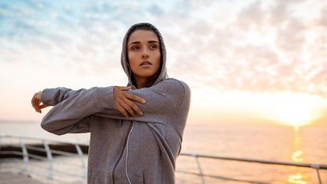 Femme s’étirant en hoodie au bord de la mer au lever du soleil.