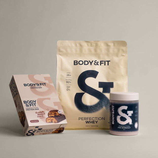 Three fitness products from BODY & FIT arranged on a neutral light gray background: a large stand-up pouch of Perfection Whey, a chocolate-flavored protein bar box, and a small supplement jar.
